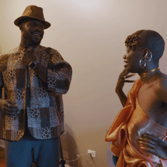 A Still from The Dilemma of Desire. Two people are standing in a room facing each other. The one to the left wears a wide-brimmed hat and a patterned jacket. The person on the right wears a shiny orange, one-shoulder top with dangly silver earrings.