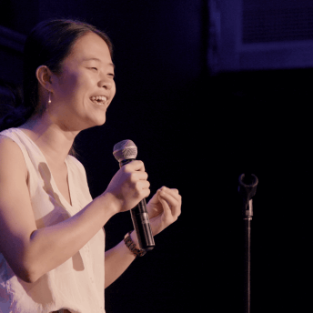 A still from The Dilemma of Desire. A person in a white tank top holds a microphone to their mouth.