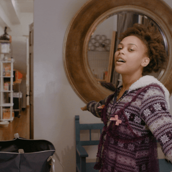 A still from The Dilemma of Desire. A person stands in an apartment with their arms held out. They are angled away from the camera, wearing a purple and white sweatshirt with a pattern on it, and a sheet tank top over top.