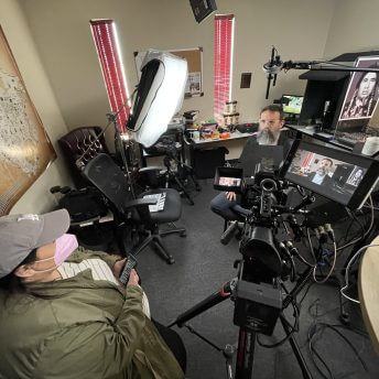 Bad Press co-director Rebecca Landsberry-Baker interviews Indigenous journalist Jerrad Moore in his Mvskoke Media office. Landsberry-Baker is wearing a gray hat and green coat and holding a cell phone with interview questions. Jerrad Moore is pictured on the camera and in-person in the photo with a long, gray beard and clear glasses.