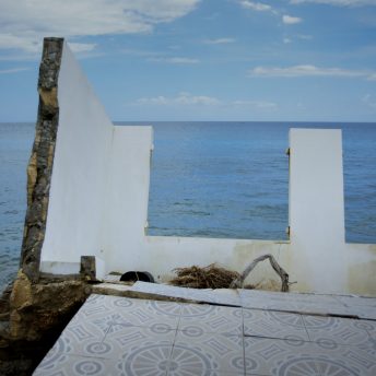 Still from Landfall. A home's wreckage on the shoreline. A wall is falling and the floor is a blue-toned mosaic. It is daytime and the sky is blue.