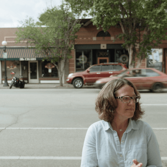 Still from Sanctuary. A woman wearing glasses stands outside and looks off-camera. She is standing across the street from a row of shops.