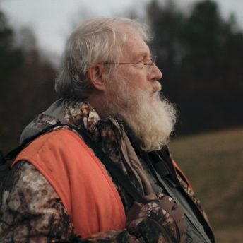 Still from One Shot One Kill. A man is standing outside, wearing a camoflauge jacket and orange vest with a gun strapped over his right shoulder.