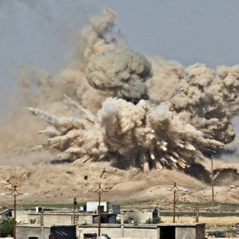 Still from Maryam Ebrahimi: 2021 Chicken & Egg Award Recipient. A large cloud of dust and smoke from an explosion in a desert. There are concrete buildings in the foreground.