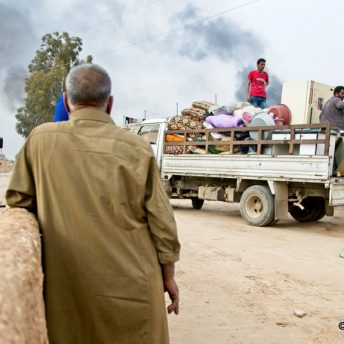 Still from Maryam Ebrahimi: 2021 Chicken & Egg Award Recipient. There are two men in a pickup truck filled with household items. Another man and woman are loading a piece of furniture onto the back of the truck. A woman with a camera is filming them. She is standing next to a man and their backs are to the camera. There is smoke billowing in the sky, and they are surrounded by sand and cinderblock buildings. There is a copyright symbol with the text "2021 Guillame Briquet" on the bottom right corner.