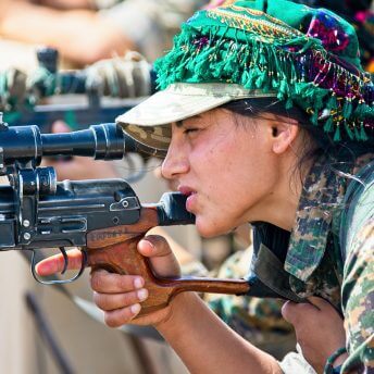 A woman in a military uniform and colorful headdress gazes into the scope of a gun leaning on a cement barrier. There are other people behind her doing the same thing.
