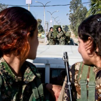 Two women in military uniforms look behind them towards two more women in uniform standing in the back of a truck.