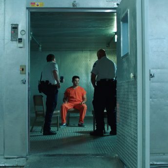 Two prison guards talk to a man in a prison uniform. They are standing in front of him, while he is sitting on a chair.