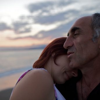 Still from The Domino Effect. A close up of woman leaning her head on a man's shoulder. His eyes are closed, and she is slightly smiling, with her face hidden in his neck. They are standing in front of water.