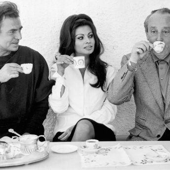 Three people drinking from tiny cups. The two people on the left are holding their cups in front of them while watching the person on the right sip from the tiny cup.