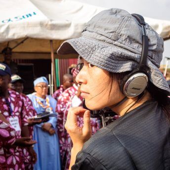 Production still of Jialing Zhang. She is wearing a wide-brimmed hat and headphones. She is pictured from the back, looking to the side and out of the frame. Behind her, slightly focused, there is a group of men in brightly-colored and patterned clothes.