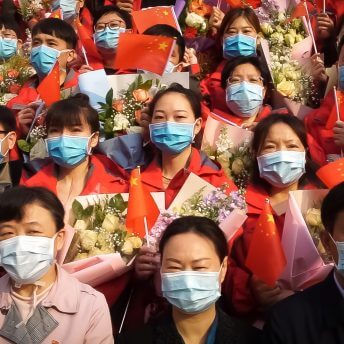 Still from In the Same Breath, a film produced by Jialing Zhang. Group portrait of Chinese men and women who are standing shoulder to shoulder in stadium-style rows. They are holding Chinese flags and some women are holding bouquets of flowers. They are all wearing blue surgical masks.