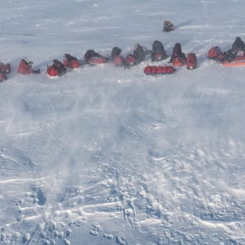 Bird's eye view of approximately 20 people in winter gear sitting and laying in a horizontal line across the snow.