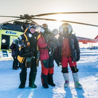 Three women in winter gear in the snow in front of a grounded helicopter, facing forward with the sun shining behind them.