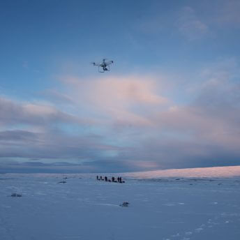 Snowy flat landscape with a drone flying in the peach and blue sky with some clouds and a group of people in the distance.