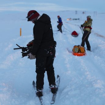 Three women in winter gear in the snow, with the closest person looking down at their camera and the others pulling supplies.