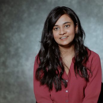 A woman with long hair and a red shirt is sitting in a set with a gray background.
