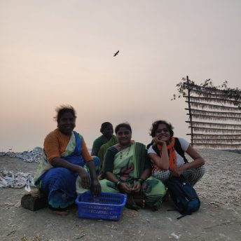 Production shot of Against the Tide. Three women sitting on the sand with baskets and with Director Sarvnik Kaur smiling.