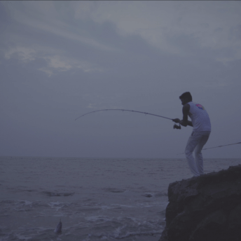 Still of Against the Tide. A man is fishing on the shore, standing on a big rock, the sun is setting and looks purple.