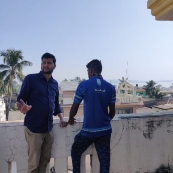 Still of Against the Tide. Two men are standing on a balcony, one of them looks at the landscape, and the other one looks in the direction of the camera. It is sunny.