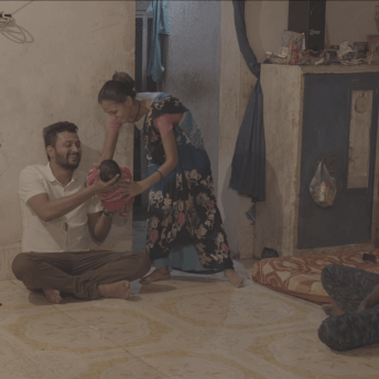 Still of Against the Tide. Inside of a house a man is sat on the floor while a woman places a baby in his arms. He smiles happily. Another man is also sat on the floor, watching the scene.