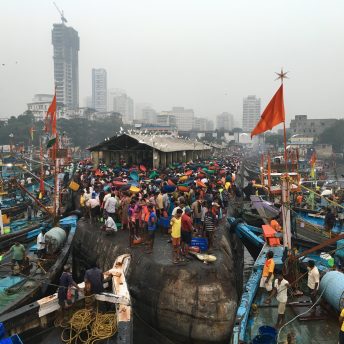 Still of Against the Tide. A shot of a crowd on the seashore, there are a lot of boats and the city is in the background.