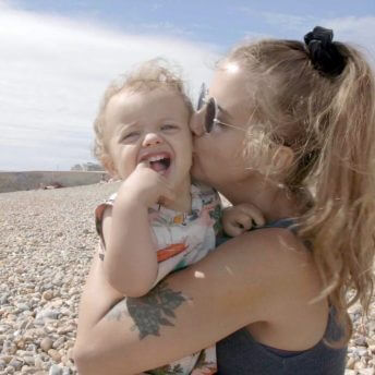 Still from Is There Anybody Out There? Ella Glendining, a blonde woman, holds her infant son River in her arms and is kissing his cheek. River's mouth is wide open and he has some fingers inside his mouth.