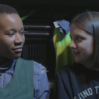 Still from Is There Anybody Out There? Ella Glednining, a blonde woman, is pictured from the chest up. She is side-by-side with another person and they both have their heads turned, looking at each other.