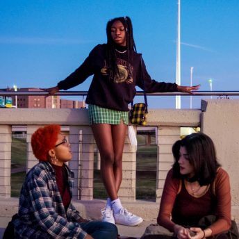 Still from Mija. Three women are on a bridge, two are sitting on the curb and one is standing against the railing.
