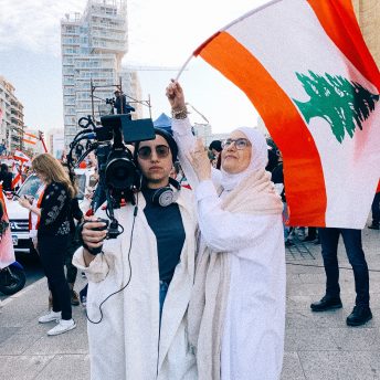 Still from Q. Filmmaker Jude Chehab is wearing sunglasses and is standing outside holding a camera over her shoulder standing next to a woman wearing glasses and a hijab, waving the flag of Lebanon, which is red and white with a green pine tree in the center.