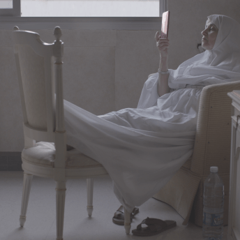 Still from Q. A woman in a hijab is sitting in a bedroom, her feet are propped on a chair and she is reading something she is holding out in front of her with her right hand.