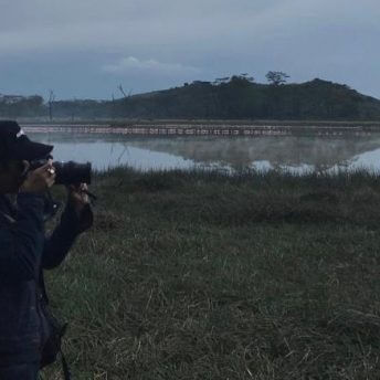 Production still of Meena Nanji looking into a camera. She stands in the middle of a field with a large pond in the center.