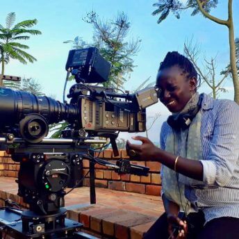 Zippy Kimundu is looking into the scope of a large camera on a tripod.