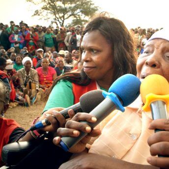 Still from Testament. An older woman speaks into three microphones being held to her mouth. Two other women, stand to the side of her, facing the camera. In the background, many people are sitting in a field looking at the three women.