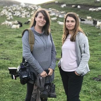 A production still from The Last Nomads. Two people stand in a field looking directly at the camera. Behind them are a patch of green grass and many sheep roaming around. One of them has a large digital camera lung over their shoulder and is holding additional camera equipment.