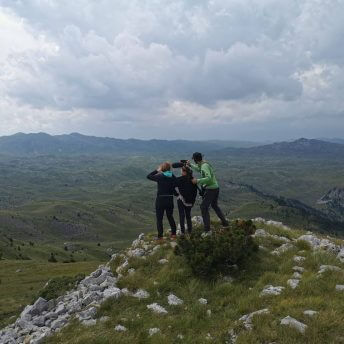 A production still from The Last Nomads. Three people stand at the peak of a green and rocky mountain. More mountains populate the horizon behind them. They stand with their backs facing the camera. One person is holding a boom.