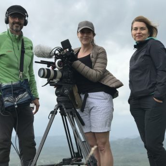 A production still from The Last Nomads. Three people stand on top of a green hill and look directly at the camera below. They are holding audiovisual recording equipment.