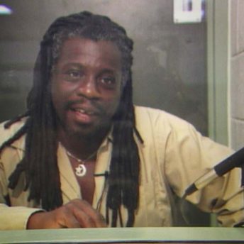 Archive photograph of Dr. Mutulu Shakur. He is talking from behind a glass screen.