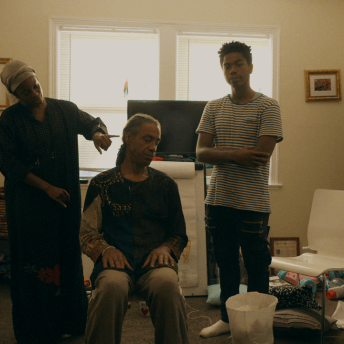 In a family room, a woman is showing how to do acupuncture to a man that is sitting on a chair. A young man is standing next to them and looks directly at the camera.