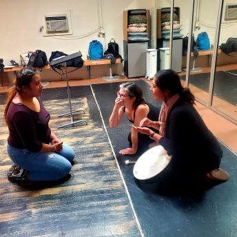 Production still from Fruits of Labor. Film protagonist, Ashley, is sitting across from film director, Emily Cohen Ibanez, and another woman who has a hand drum leaning against her knees.