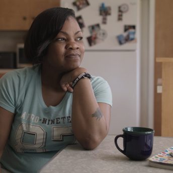 Still from Since I Been Down. A woman is sitting in a kitchen, leaning her right arm on a table and looking away from the camera.