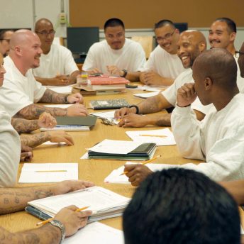 Still from Since I Been Down. A group of men in prison is sitting at the table, they have papers and pencils. Some of them smile.