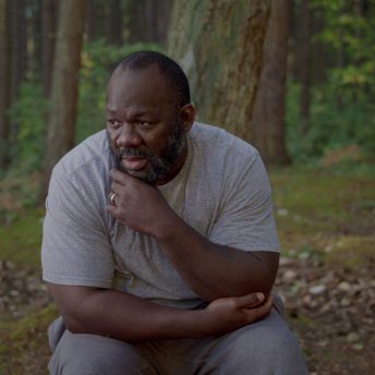 A man is sitting in a trunk in a forest. He reclines his left arm on his lap and uses the other arm to hold his face. He looks away from the camera.