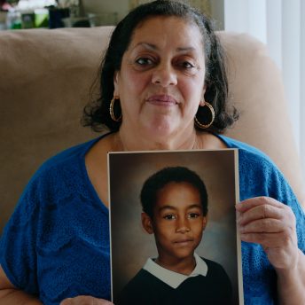 A woman with a blue shirt holds the portrait of a young boy with both of her hands. She looks directly at the camera and is sitting in a sofa.