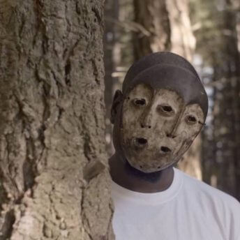 Still from Since I Been Down. A person wearing a mask peeks behind a tree.