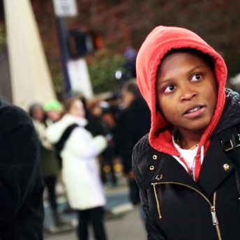 Still from Unapologetic. A person is standing outside in a black jacket with a red hood pulled up and surrounded by others in winter clothing.