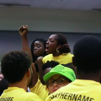 Still from Unapologetic. People gathered, wearing yellow shirts, and one of them has their fist up in the air. A woman stands out from the crowd.