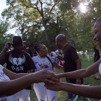 Still form Unapologetic. Two women stand outside, smiling and holding hands surrounded by others.