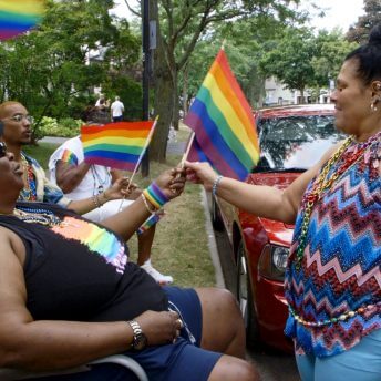 Still from And So I Stayed. Three people are sitting on chairs, holding LGBTQ+ flags, and a woman is standing in front of one of them and grabbing one of the flags. There is a red truck parked.
