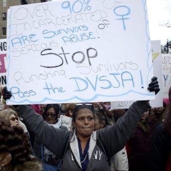 Still from And So I Stayed. In the middle of a demonstration, Kim Dadou Brown is holding a poster that says "Over 90% of incarcerated are survivors of serious abuse! Stop punishing survivors. Pass the DVSJA!"